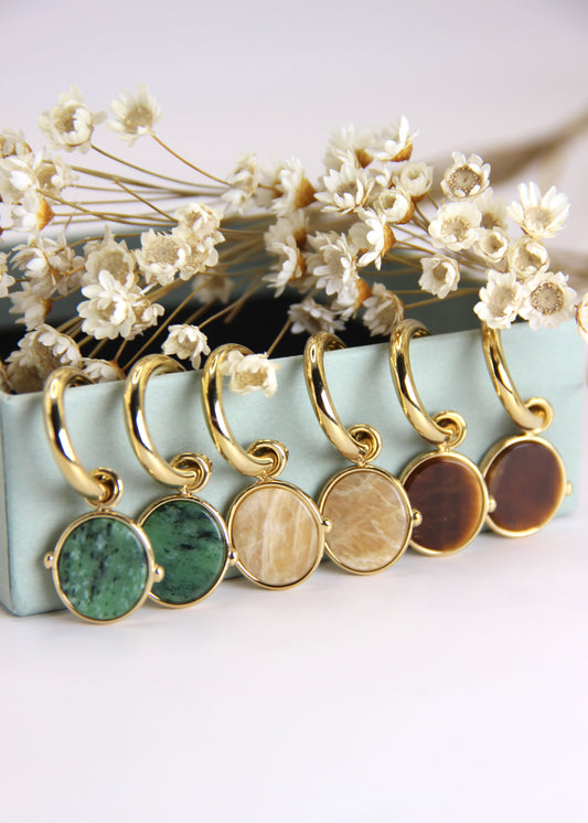 Orecchini a cerchio in acciaio placcato oro con pendente ovale in pietra dura naturale. Modello elegante con chiusura a perno, disponibile in vari colori tra cui occhio di tigre, malachite e opalite. Ogni pietra presenta leggere variazioni naturali di colore e venature.