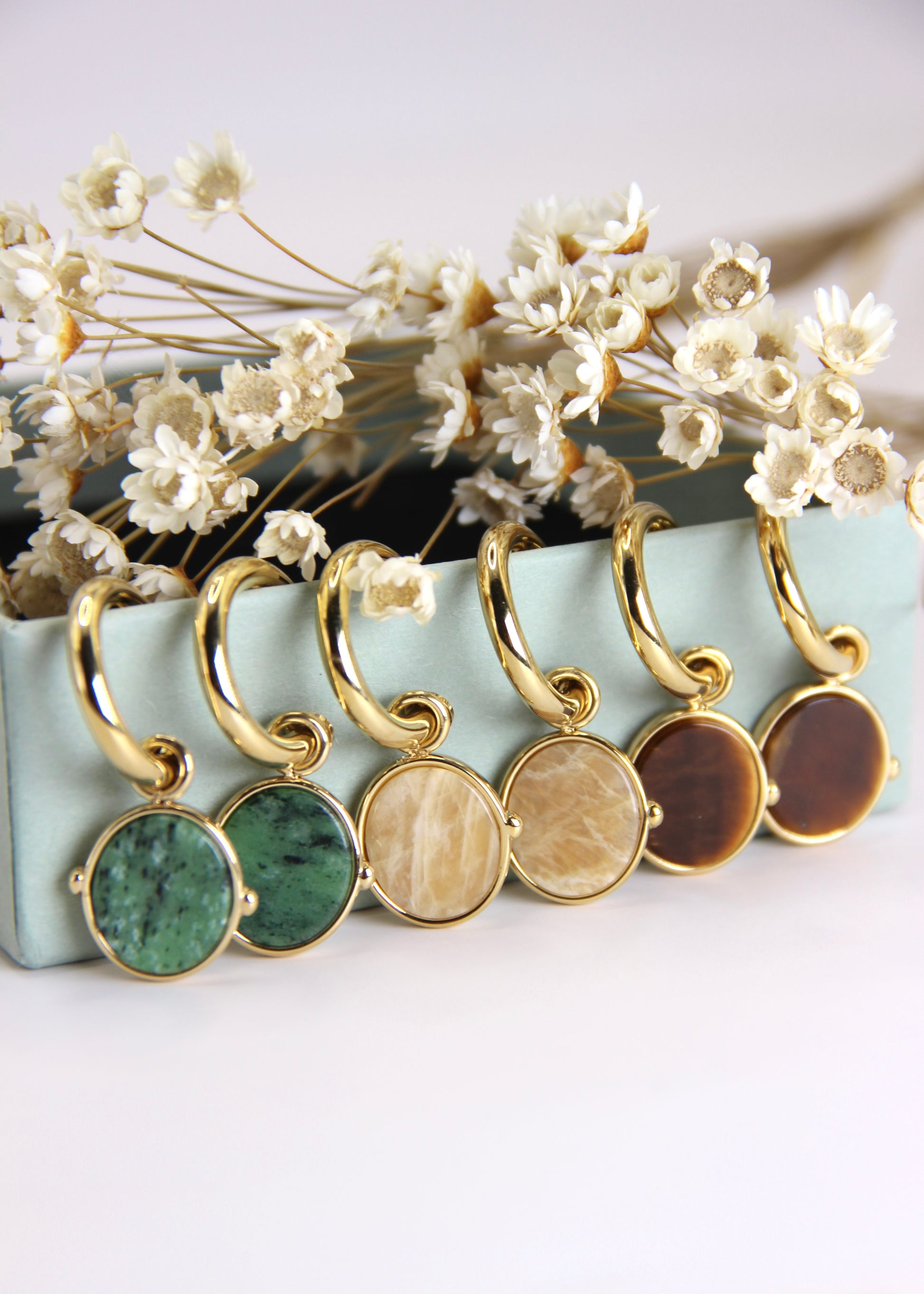 Orecchini a cerchio in acciaio placcato oro con pendente ovale in pietra dura naturale. Modello elegante con chiusura a perno, disponibile in vari colori tra cui occhio di tigre, malachite e opalite. Ogni pietra presenta leggere variazioni naturali di colore e venature.